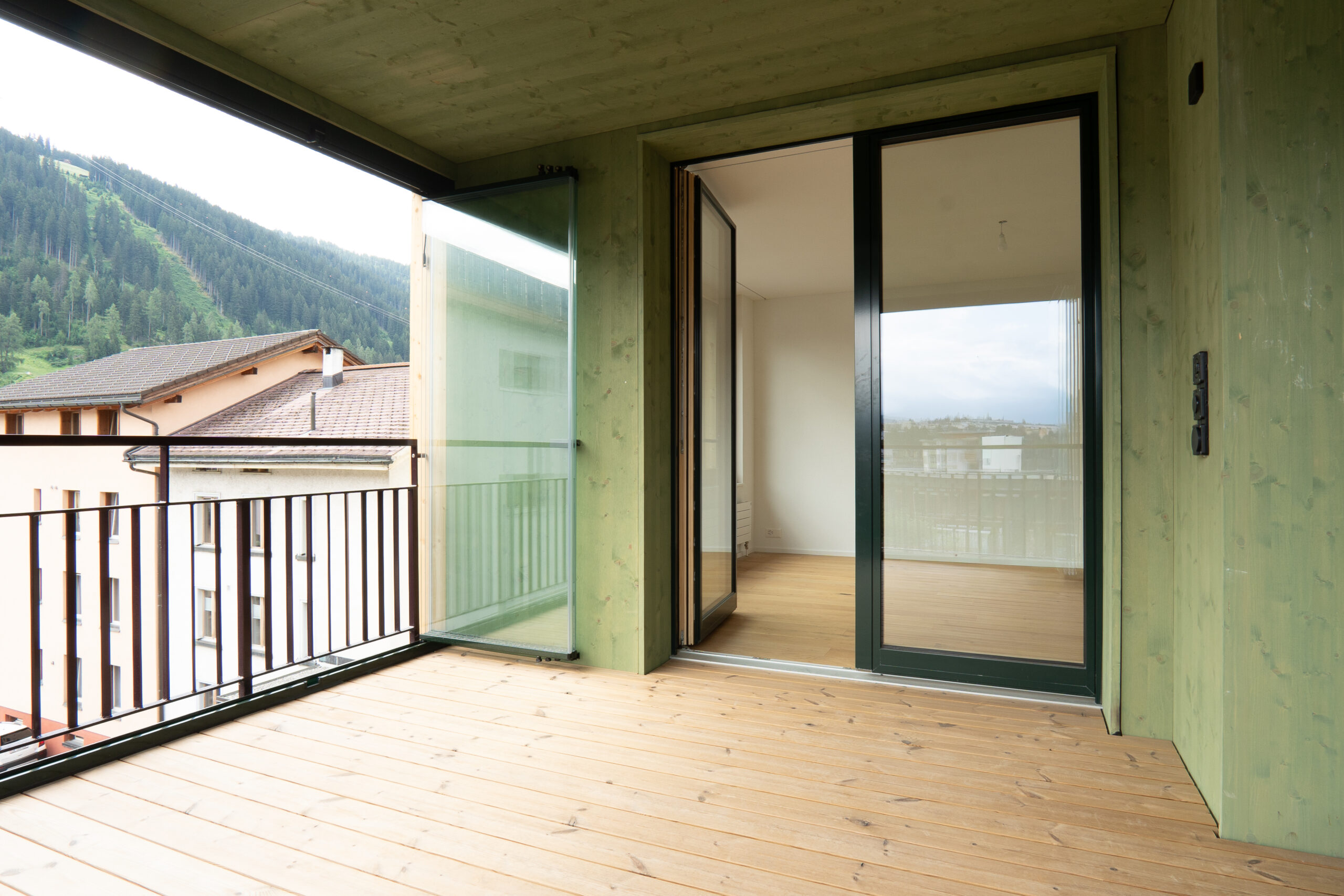 Überdachter Balkon mit Holzboden, grünlicher Holzverkleidung, Glaswindschutz und Blick auf die Berglandschaft.