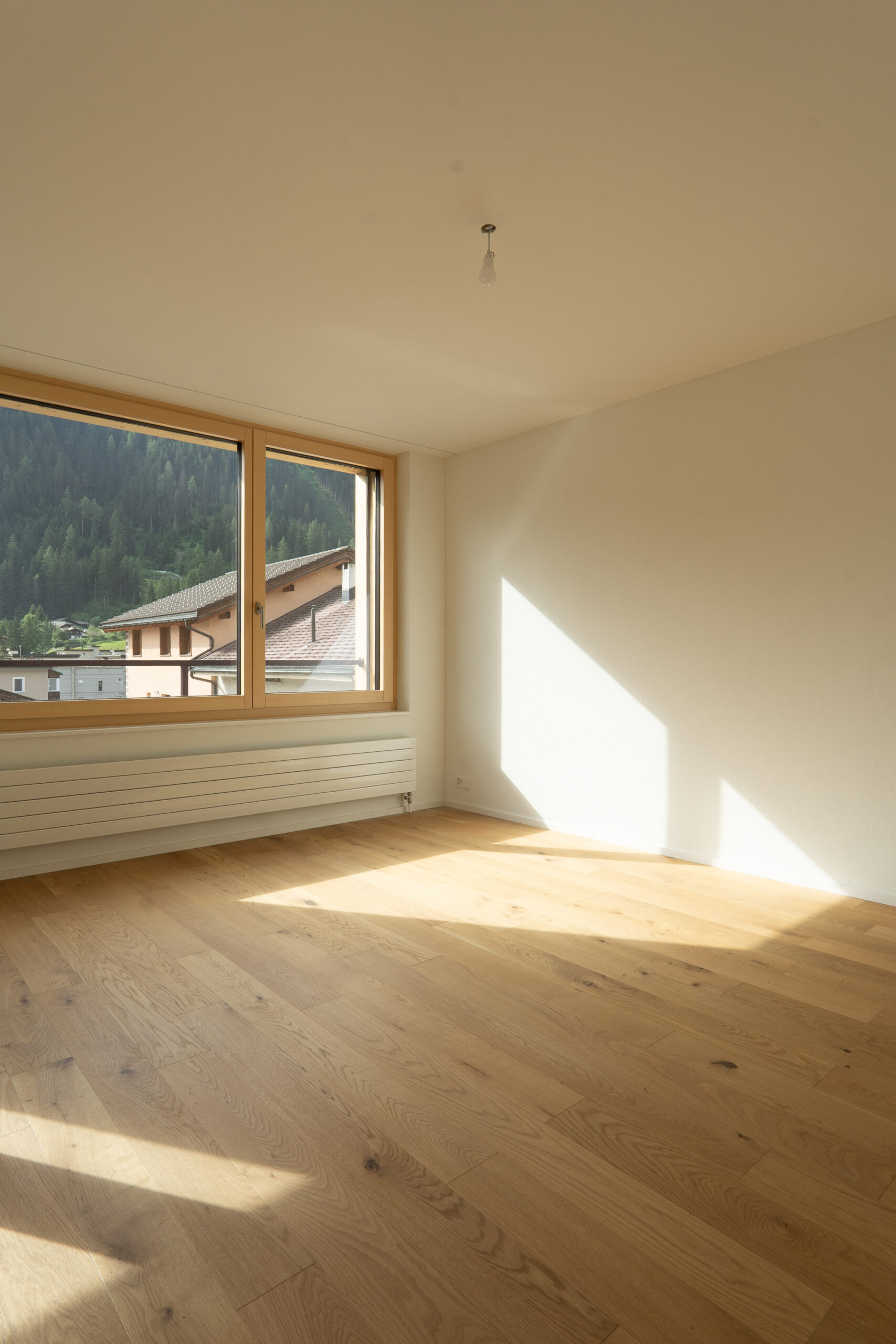 Sonnendurchfluteter Wohnraum mit natürlichem Eichenparkett, weissen Wänden und Holzfenstern mit Blick auf die Berge.