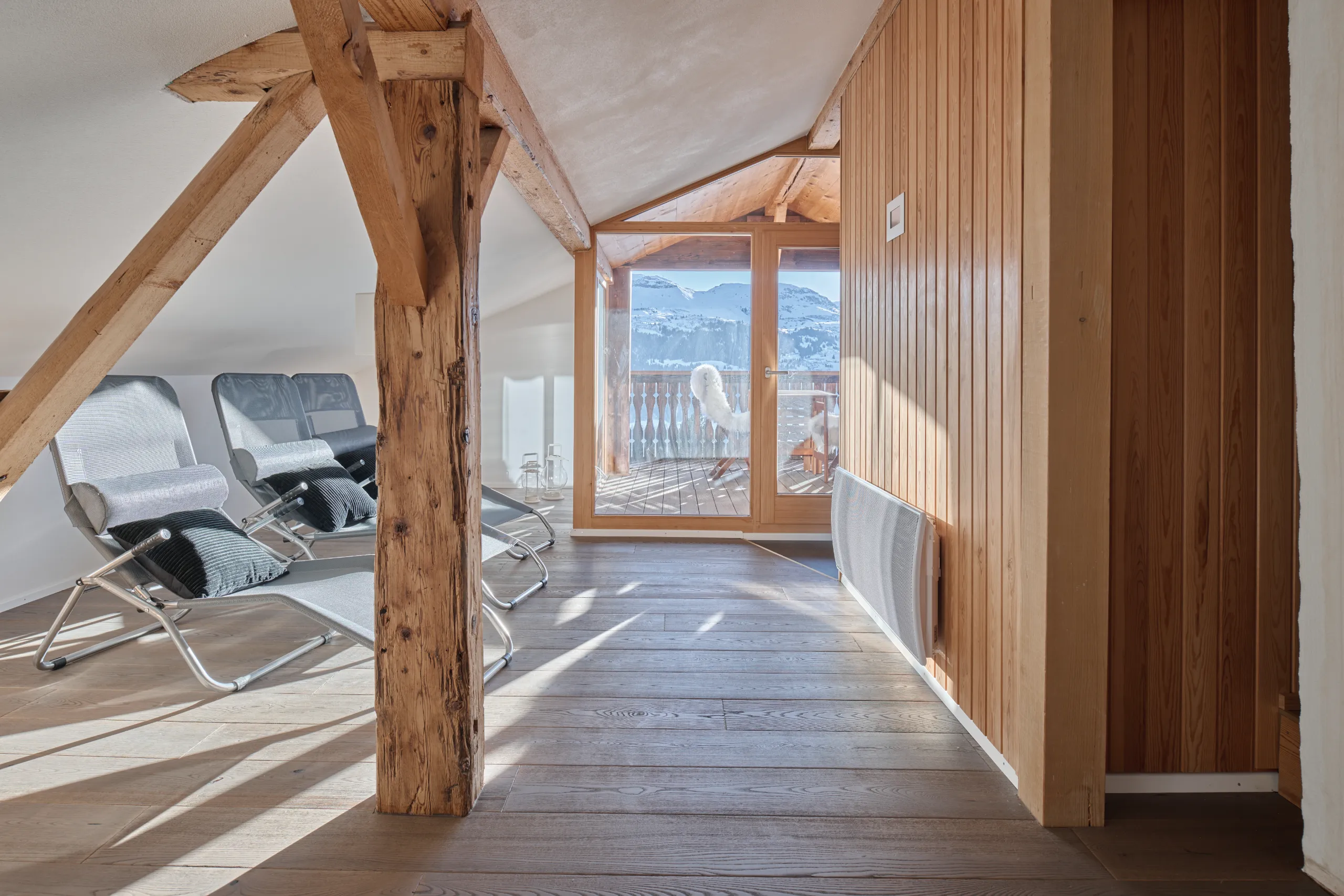 Gemütlicher Ruheraum mit sichtbaren Holzbalken, Holzverkleidung, Liegestühlen und Blick auf die verschneite Berglandschaft.