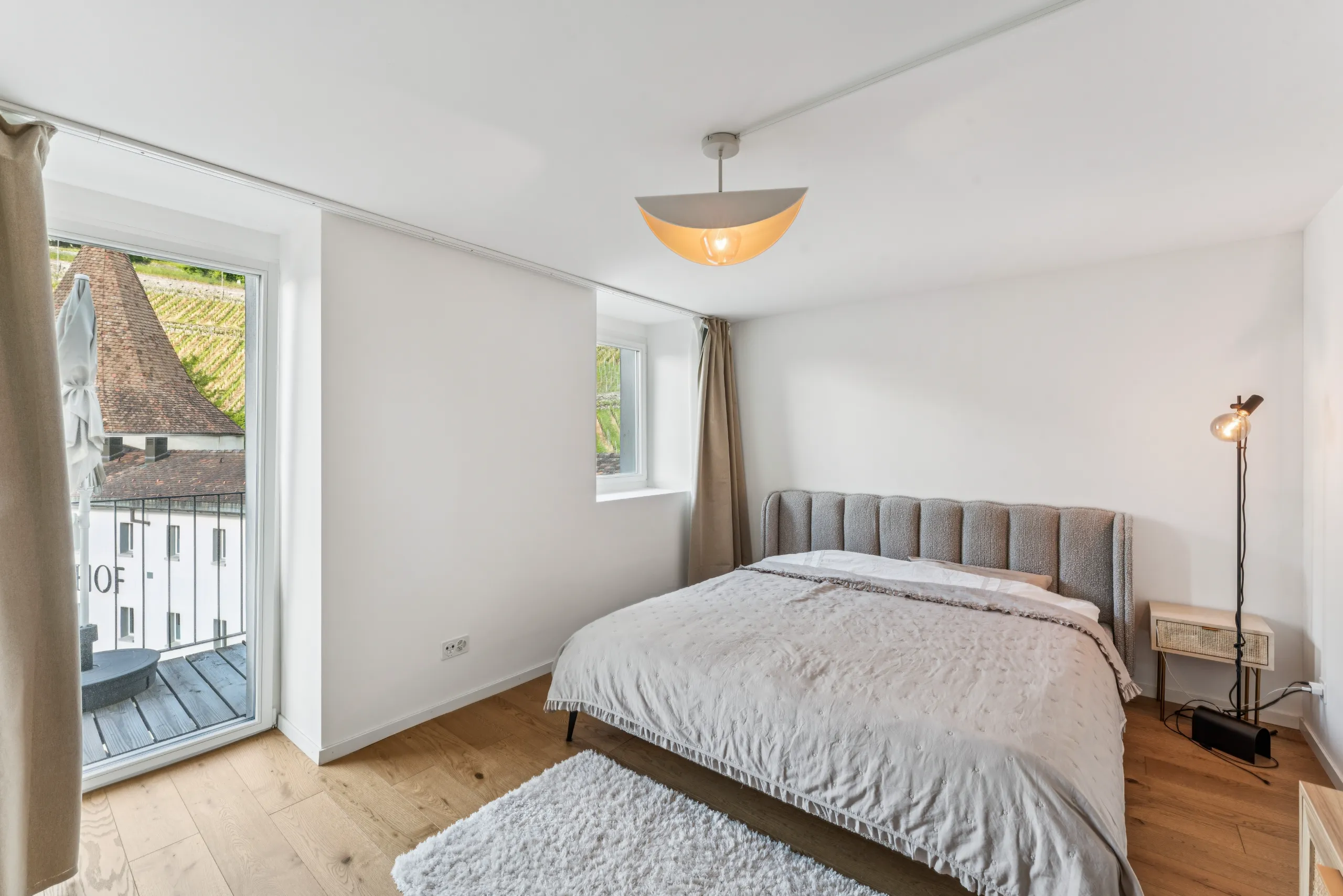 Helles Schlafzimmer mit natürlichem Eichenparkett, gemütlichem Doppelbett und grossen Fenstern mit Balkonzugang.