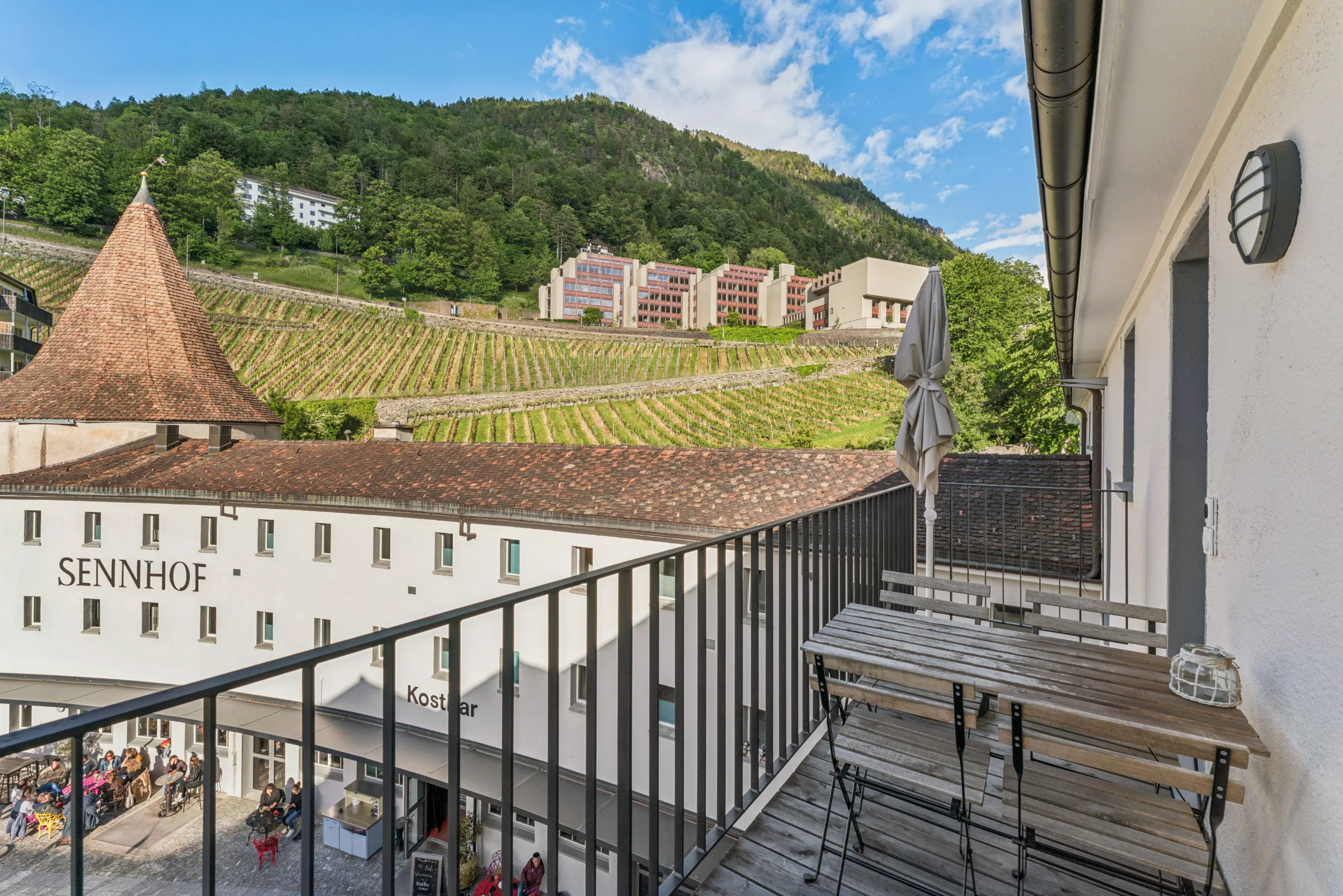 Balkon mit Holztisch und Blick auf Sennhof, Rebberg und bewaldeten Hang in Chur.