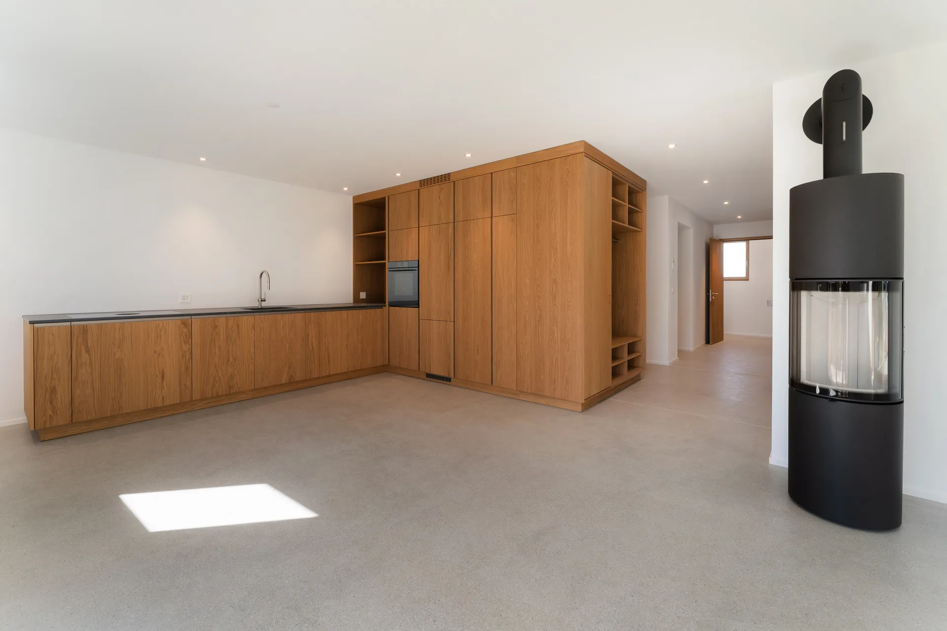 Offener Wohnbereich mit moderner Küche aus Holz, schwarzem Cheminée und hellem Bodenbelag.
