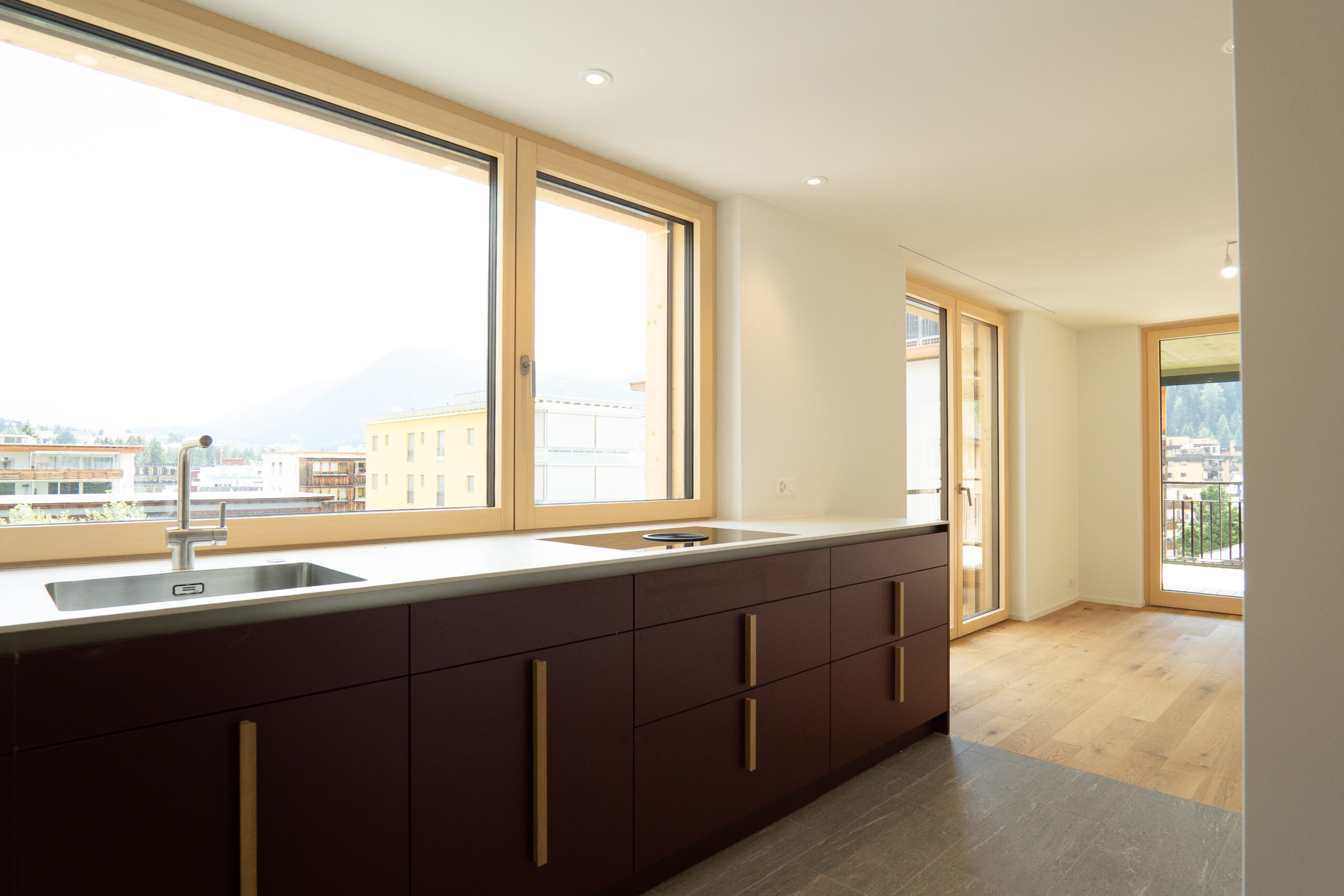 Moderne Küche mit bordeauxroten Fronten, Holzgriffen, heller Arbeitsplatte und grosszügigen Holzfenstern.
