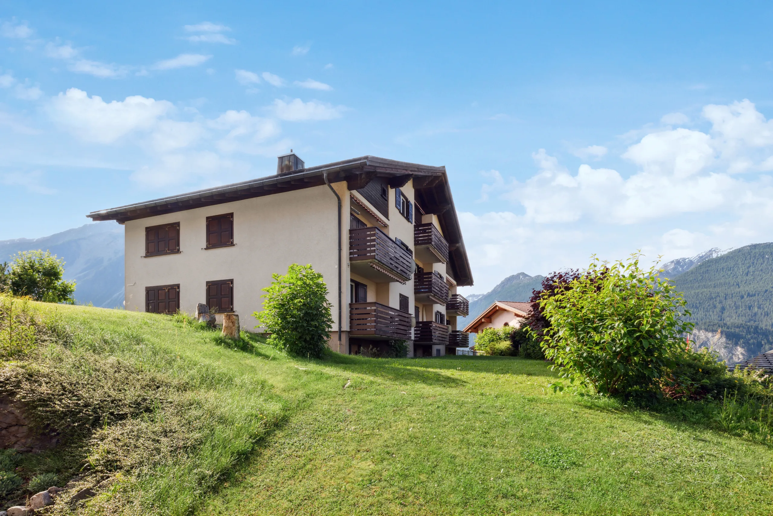 Dreistöckiges Mehrfamilienhaus mit Holzbalkonen auf grüner Wiese vor Bergkulisse in Schmitten.