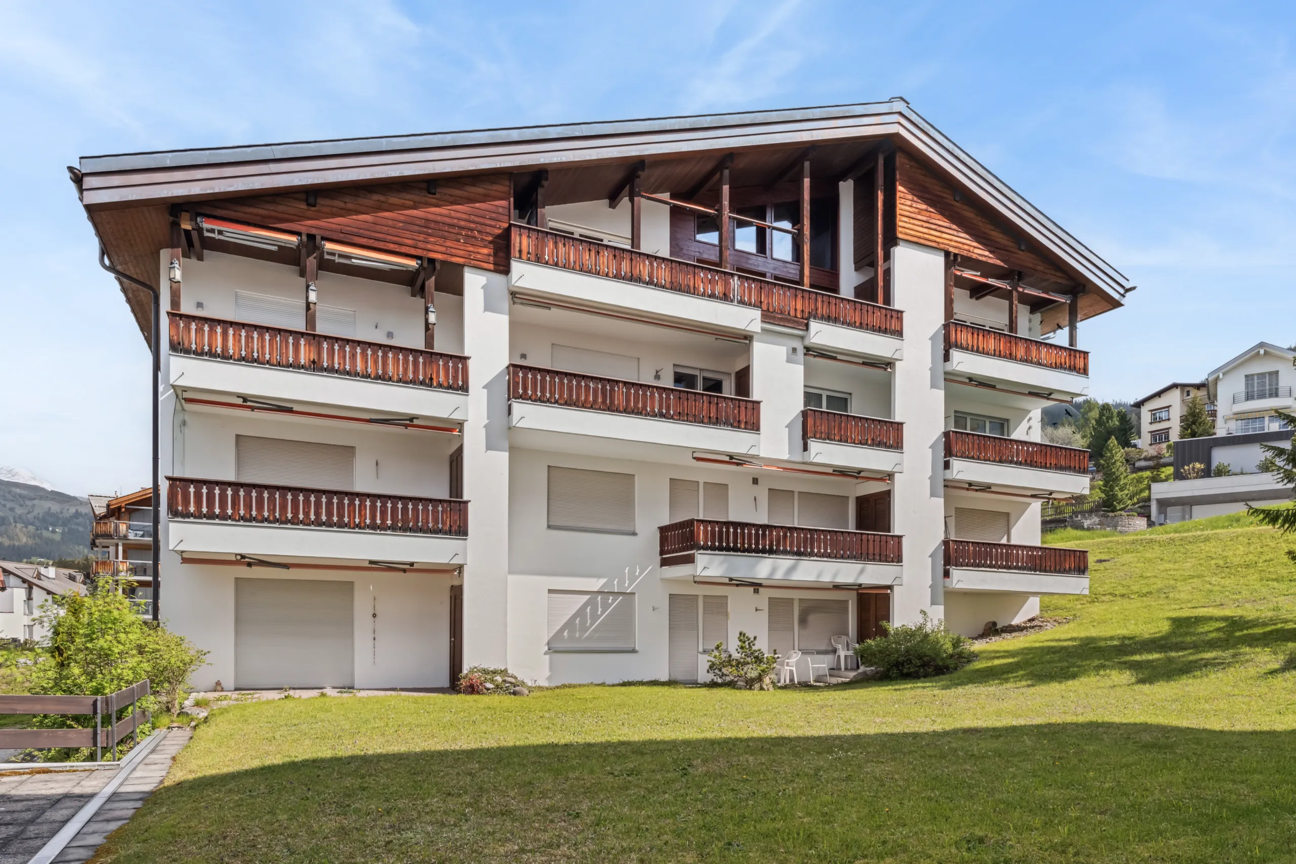 Mehrfamilienhaus in Lenzerheide mit weisser Fassade, traditionellen Holzbalkonen und gepflegter Grünanlage.