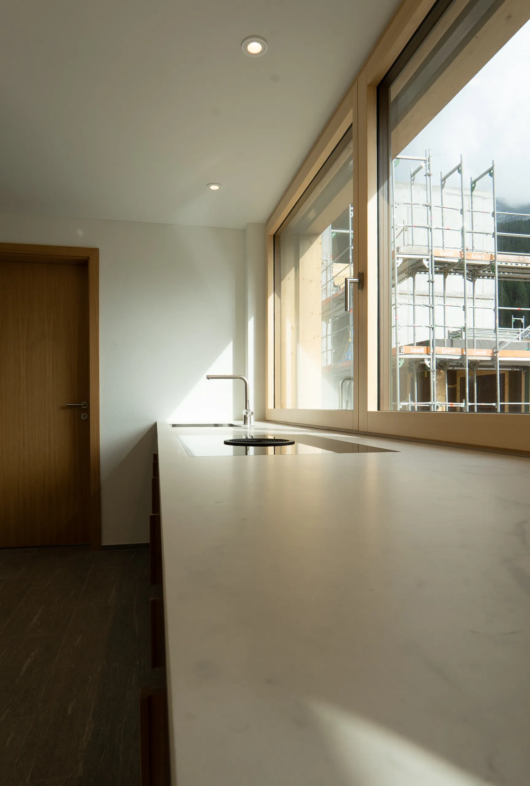 Helle Küchenarbeitsplatte mit integriertem Kochfeld, Spülbecken und grossen Holzfenstern entlang der Arbeitsfläche.