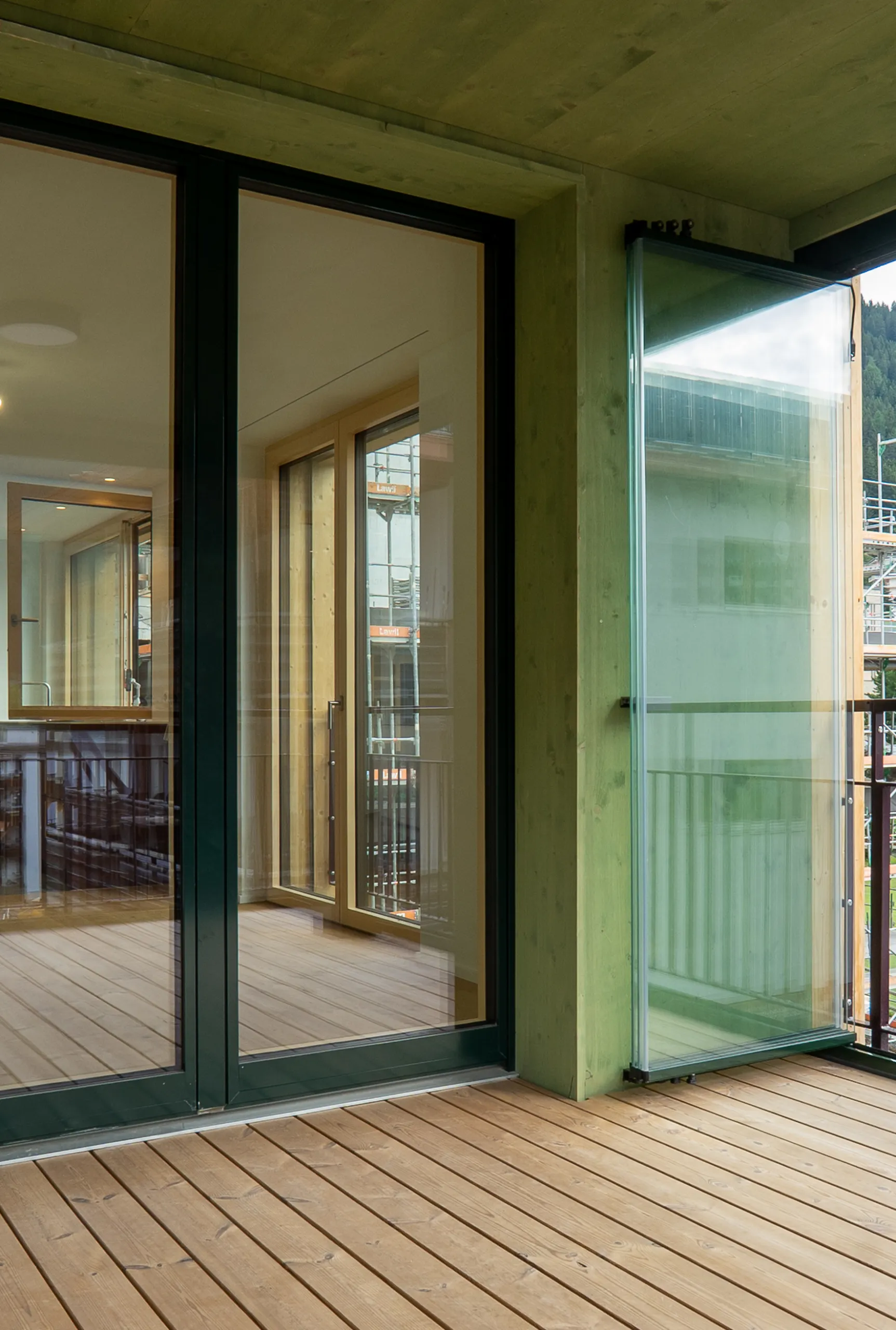 hoch-6-scaled Überdachter Balkon mit Holzdielen, grünlicher Holzverkleidung, schwarzen Fenstertüren und seitlichem Glaswindschutz.