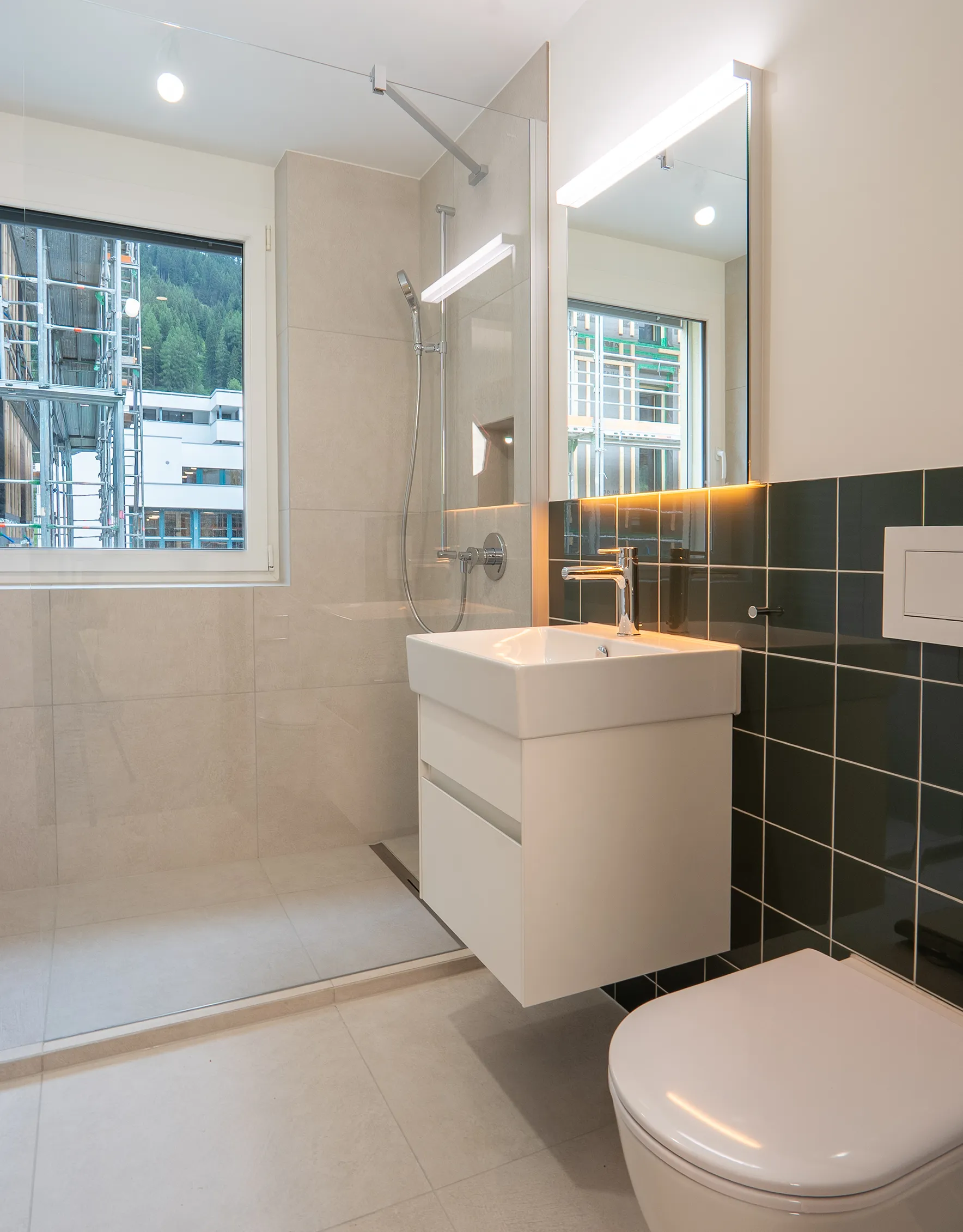 Modernes Badezimmer mit bodenebener Dusche, weissem Waschtischunterschrank, beleuchtetem Spiegel und dunkelgrünen Wandfliesen.