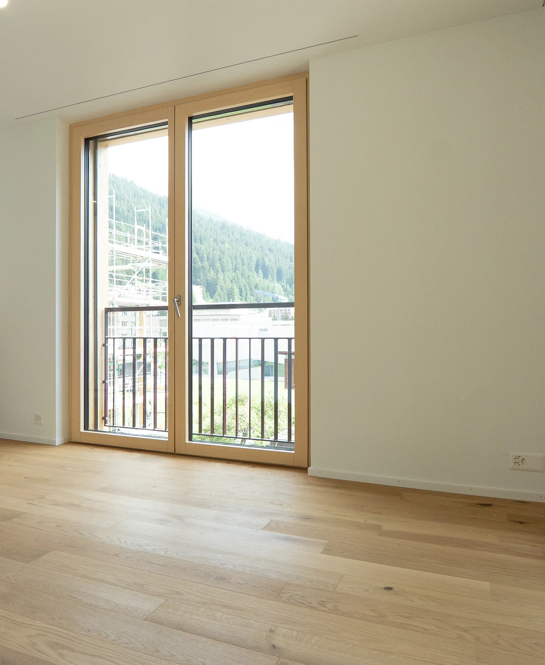 mittel-hoch-3 Heller Wohnraum mit natürlichem Eichenparkett, bodentiefen Holzfenstertüren und Blick auf die bewaldeten Berge.