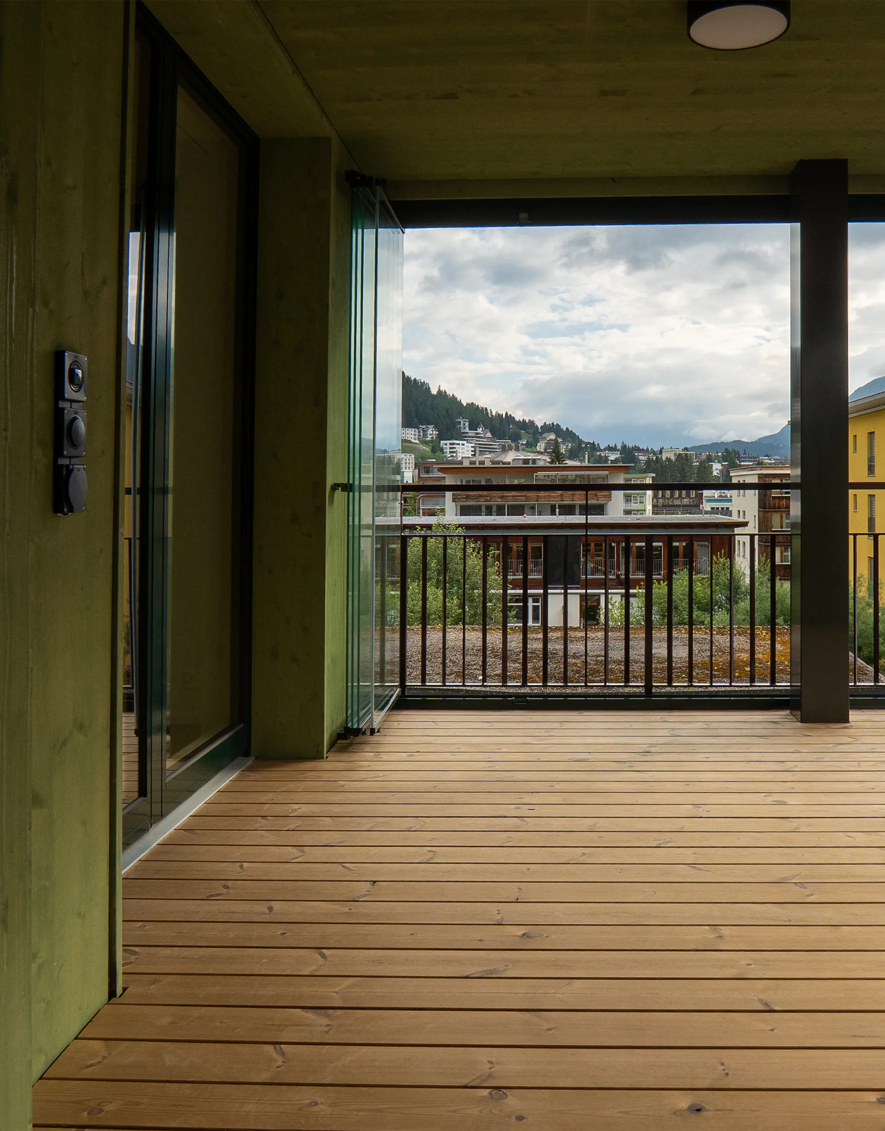 Überdachter Balkon mit Holzdielen, grünlicher Holzverkleidung, schwarzem Geländer und Ausblick auf Davos.
