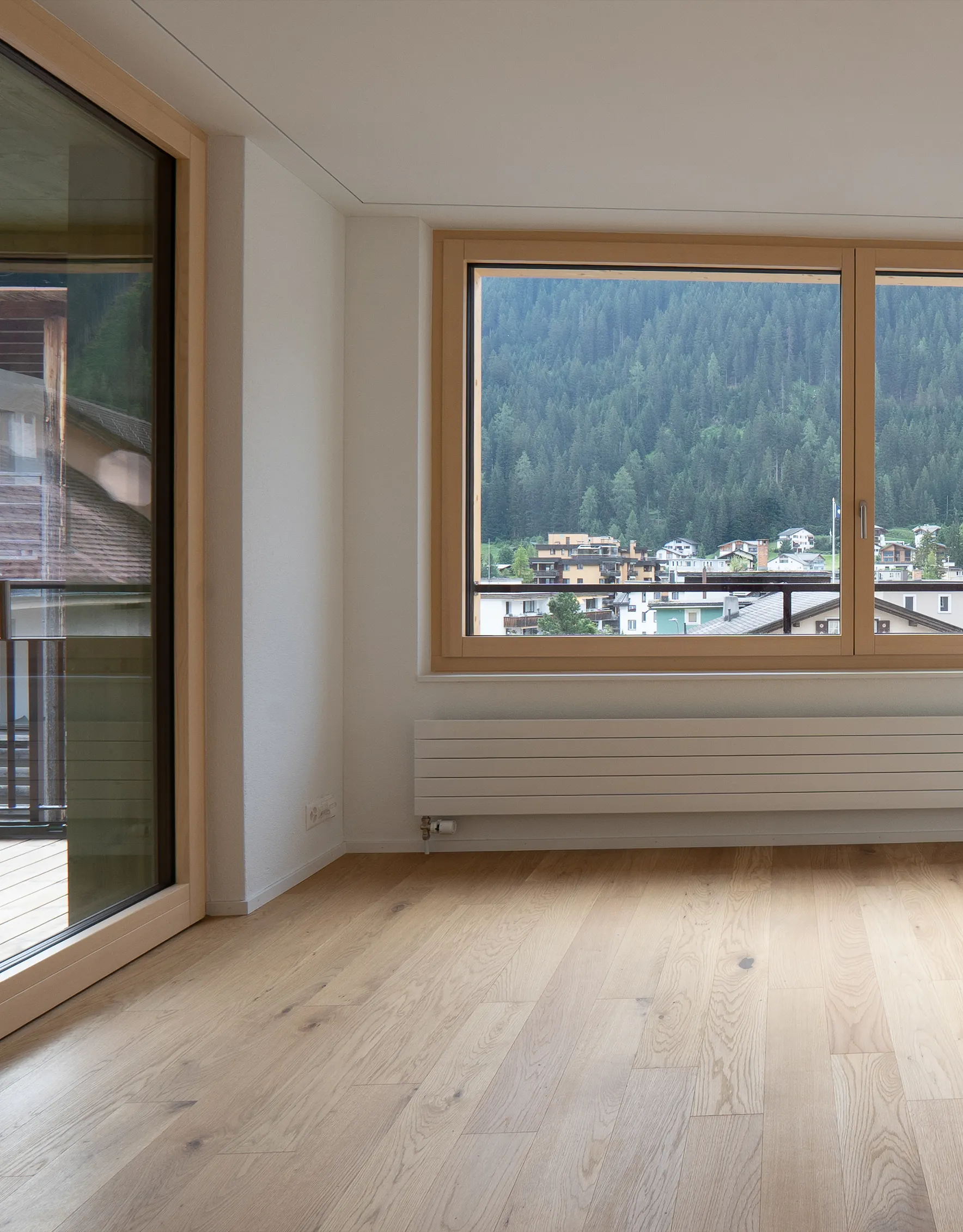 mittel-hoch-6 Heller Wohnraum mit natürlichem Eichenparkett, grossen Holzfenstern und Aussicht auf die bewaldete Berglandschaft.