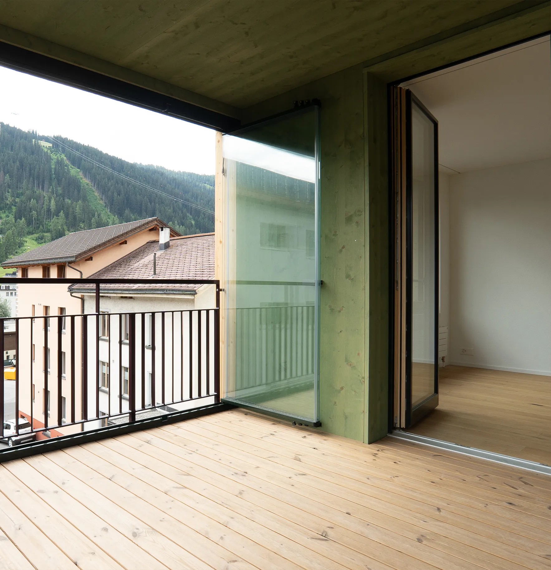 mittel-tief-3 Überdachter Balkon mit Holzdielen, grünlicher Holzverkleidung, Glaswindschutz und Aussicht auf die Berglandschaft.