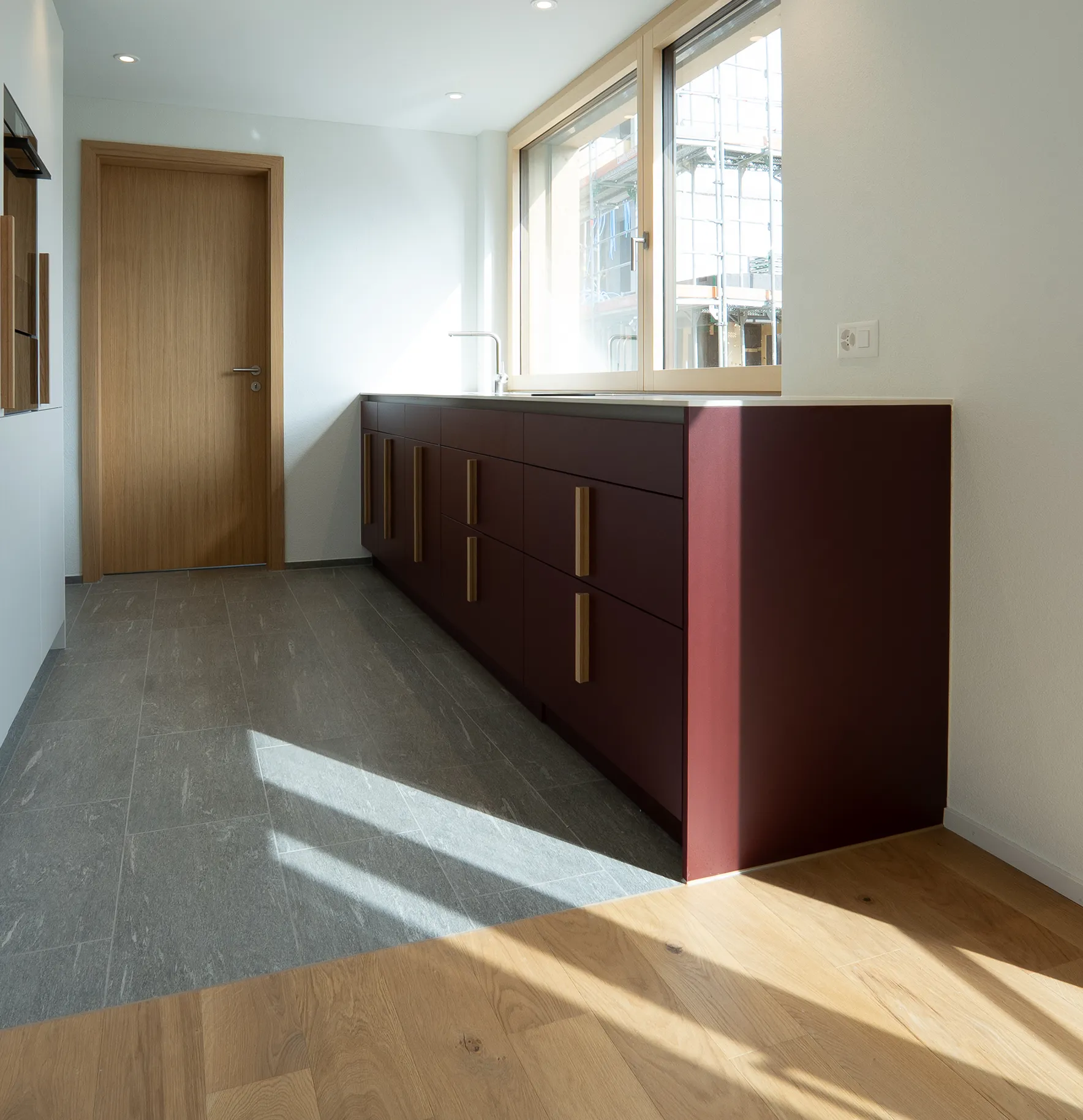 Moderne Küche mit bordeauxroten Unterschränken, langen Holzgriffen, heller Arbeitsplatte und angrenzendem Essbereich.
